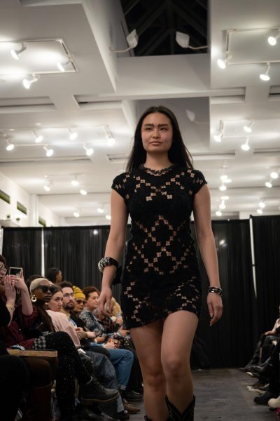 A model walks down a runway wearing a black dress with diamond patterns and one black and gray bracelet on each of their wrists.
