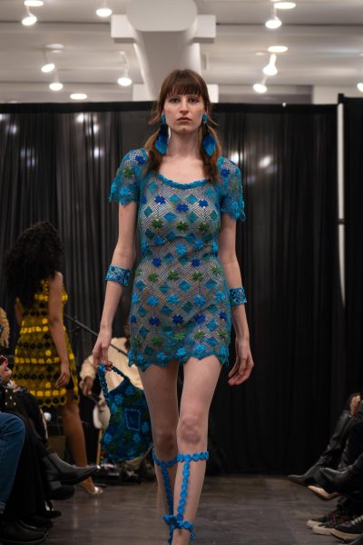 A model walks down a runway wearing a blue lace dress with blue and green floral patterns and a blue bracelet on each of their forearms.