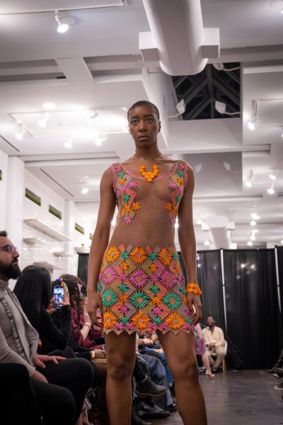 A model poses on a runway wearing a lace dress with orange, green and pink floral patterns and an orange bracelet with circles on their left hand.