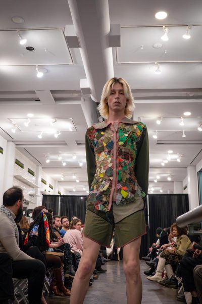 A model walks down a runway wearing a dark green shirt and dark green shorts with red linings on the borders. On the shirt is a collage of colorful pieces of cloth.