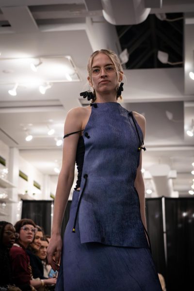 A model walks down a runway in a blue high neck top with black strings and a matching blue skirt. The model is wearing big black dangling earrings.
