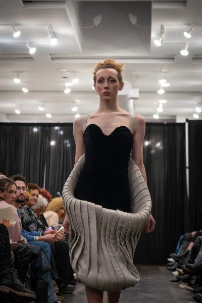 A model walks down a runway in a black dress with beige straps and a beige pleated cylinder around her upper back and lower stomach.