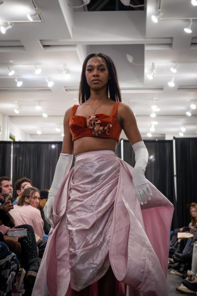 A model walks down a runway in a baby pink layered skirt and an orange top with a floral design. The model is wearing white gloves.