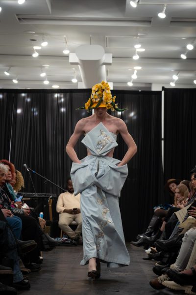 A model walks down a runway wearing a yellow flower hat and a light blue dress with protruding pockets and gold accents.