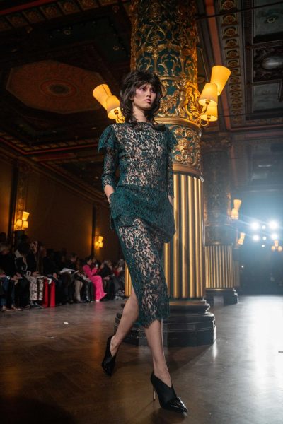 A model wearing a green lace dress struts past a golden pillar.