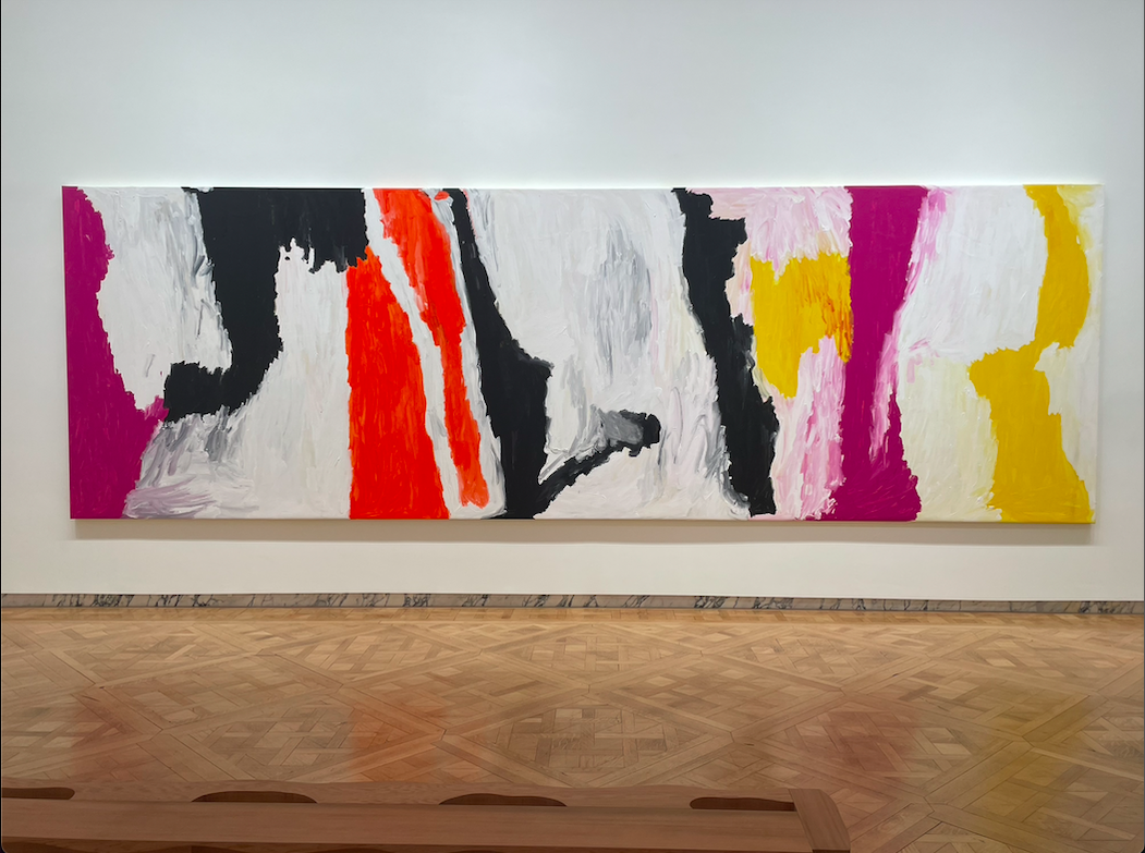 An abstract painting with white, black, yellow, pink and orange placed on a white wall.