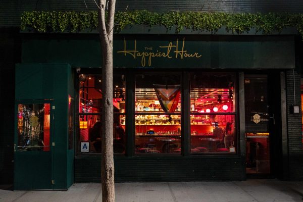 A bar illuminated with red and yellow lights is visible through the window of a brick building. The top of the building has gold lettering that says “THE Happiest Hour.”