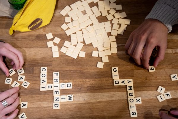 Two pairs of hands arrange small letter tiles into columns and rows of words. There is a pile of tiles on the table next to a yellow sack labeled “BANANAGRAMS.”