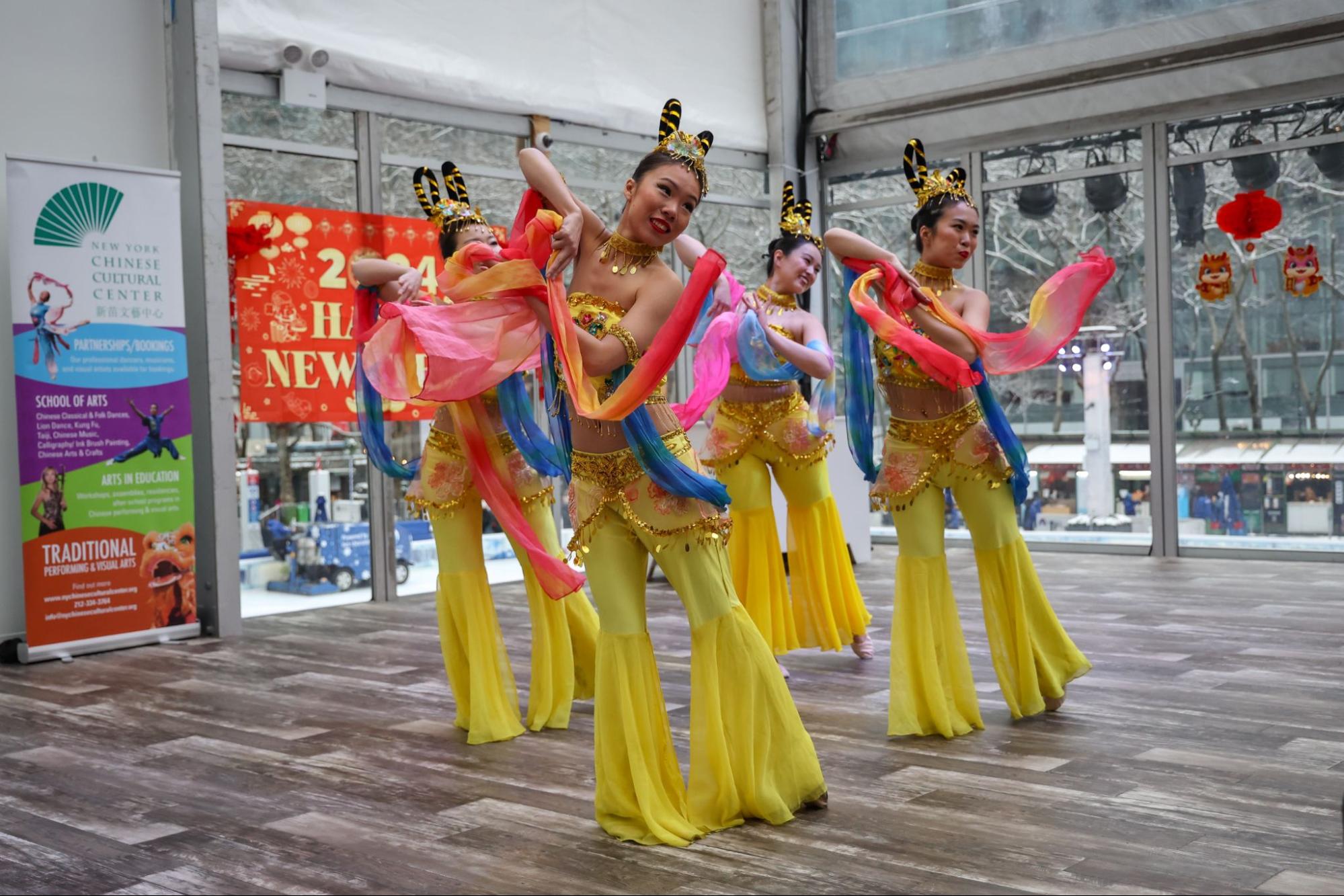 Four dancers wearing yellow outfits perform a dance in a room while holding red and blue ribbons.