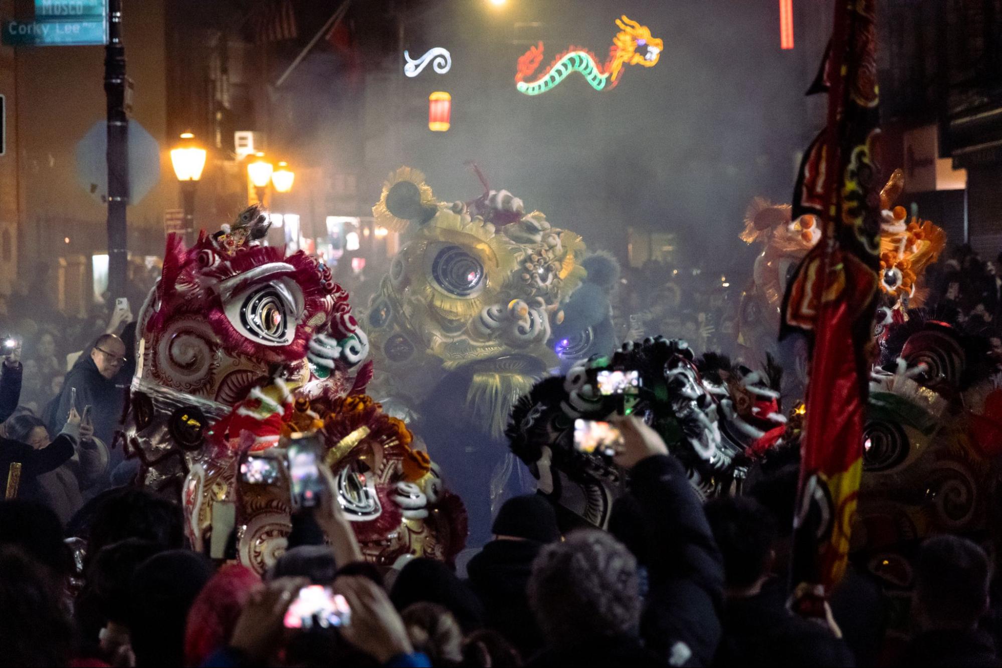 A number of lions dancing in a circle in the middle of a street amidst smoke and a crowd of audience.