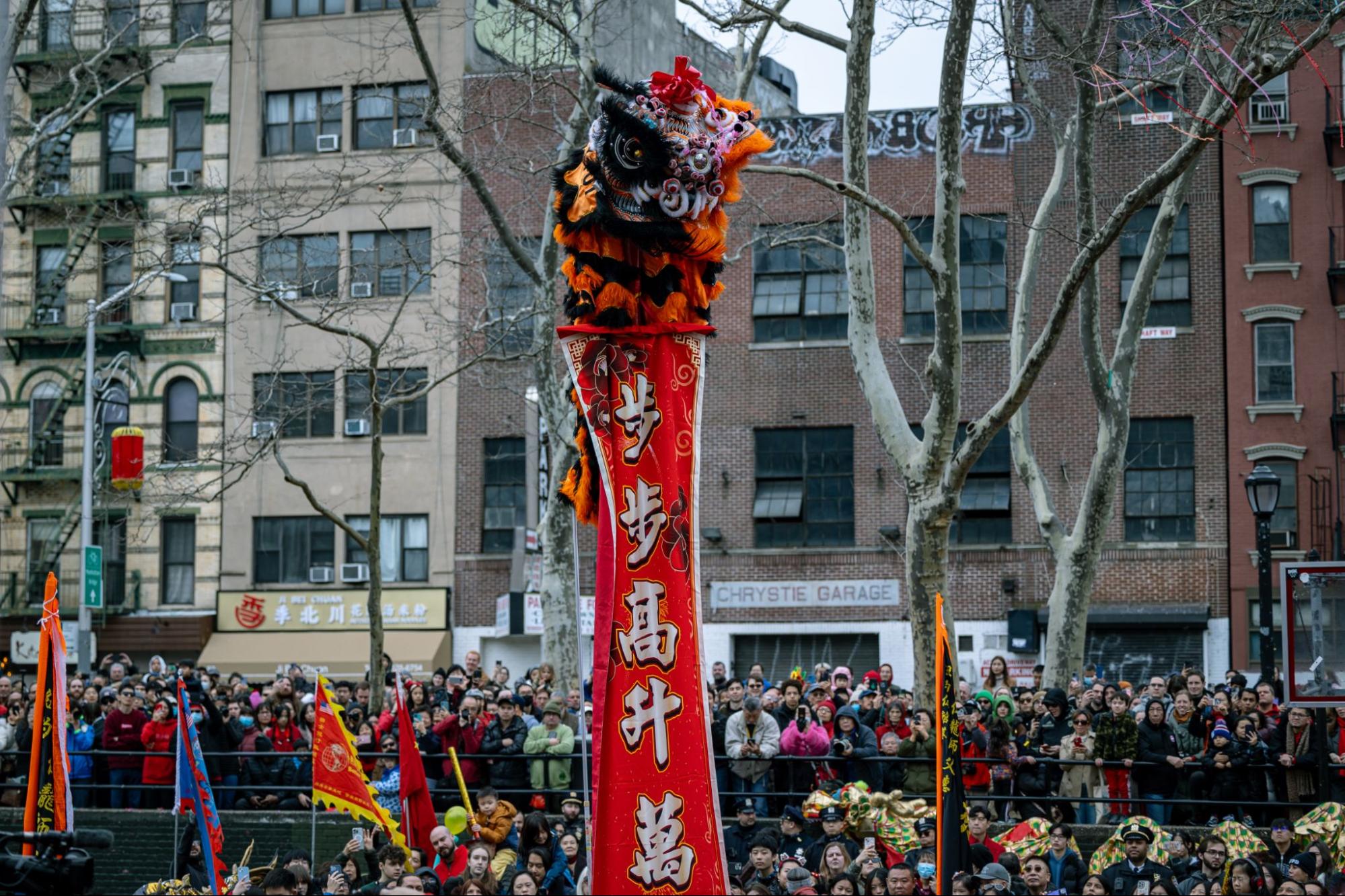 A lion dancer stands on a pole in the center of a stage. Tens of people are watching the lion dancer unravel a long red scroll with white Chinese characters.