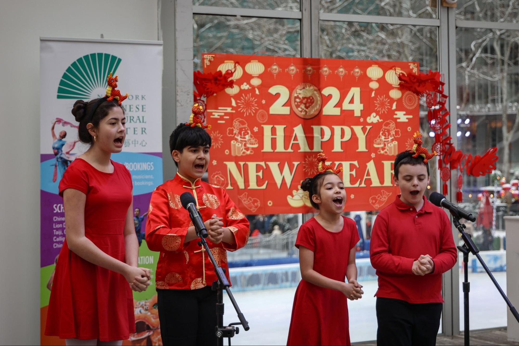 Four children wearing red outfits sing in unison towards a crowd. Behind them is a red poster that says “2024 HAPPY NEW YEAR.”