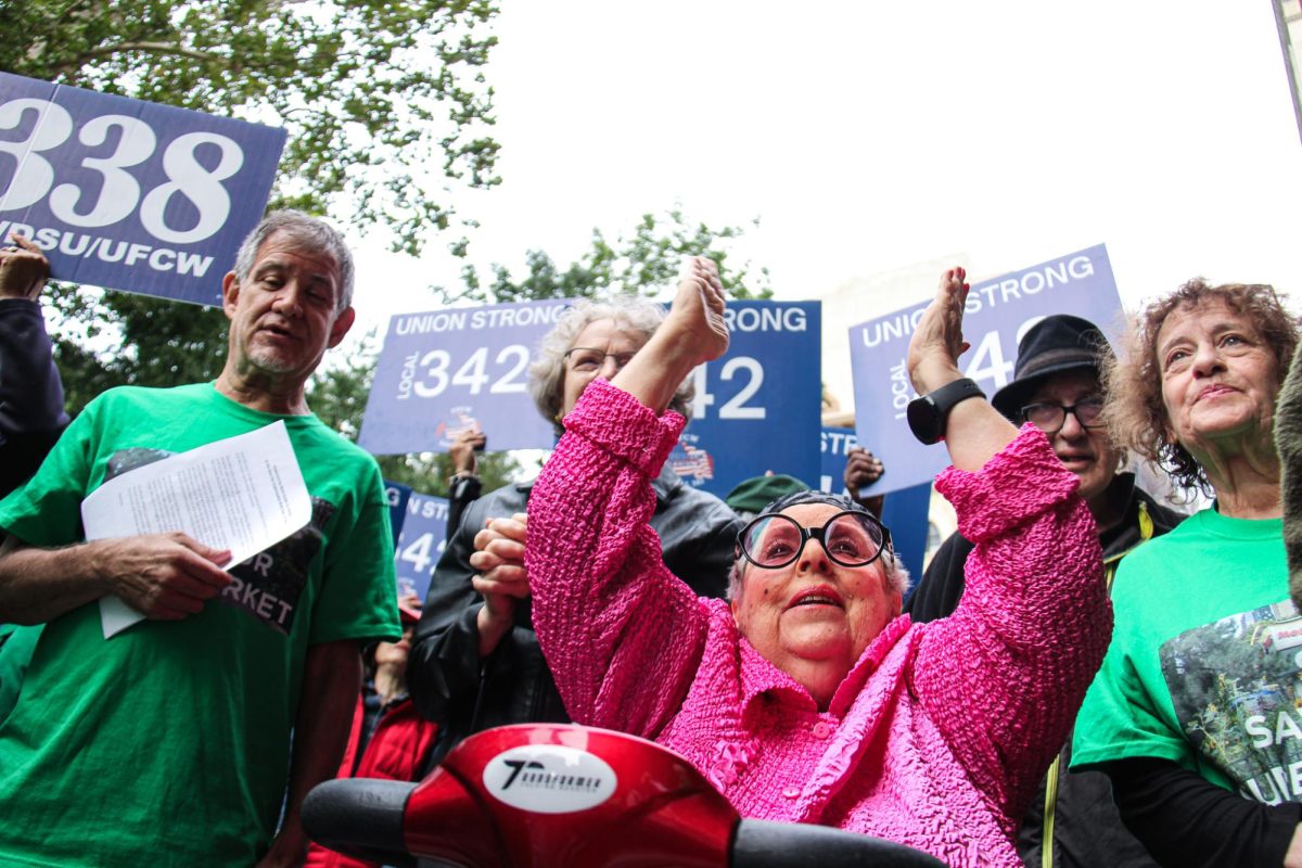 A woman in a pink jacket with black glasses, surrounded by individuals in green shirts, claps during a protest being held to save the Morton Williams supermarket. In the background, there are blue signs that read, “Union Strong Local 342.”