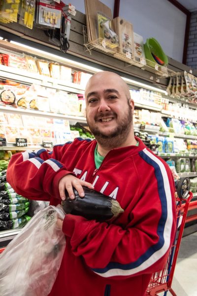 A man wearing a red jacket and a green shirt holding a plastic bag and an eggplant stands in a supermarket.