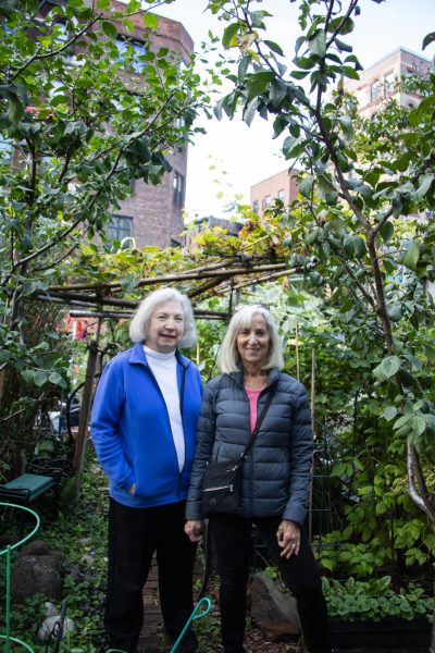 A woman wearing a white shirt and a blue jacket and a woman with a pink shirt with a blue jacket stand side by side in a garden.