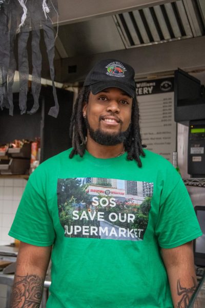 A man wearing a green t-shirt that says "S.O.S. Save Our Supermarket" and a black cap smiles in front of the camera.