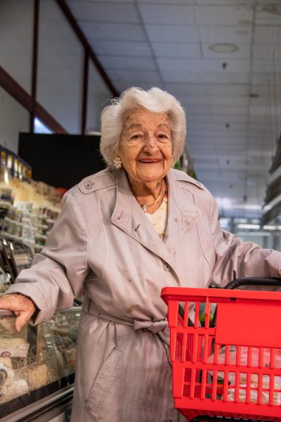 An elderly woman wearing a gray trench coat holds a red grocery basket in her left hand as she smiles at the camera.