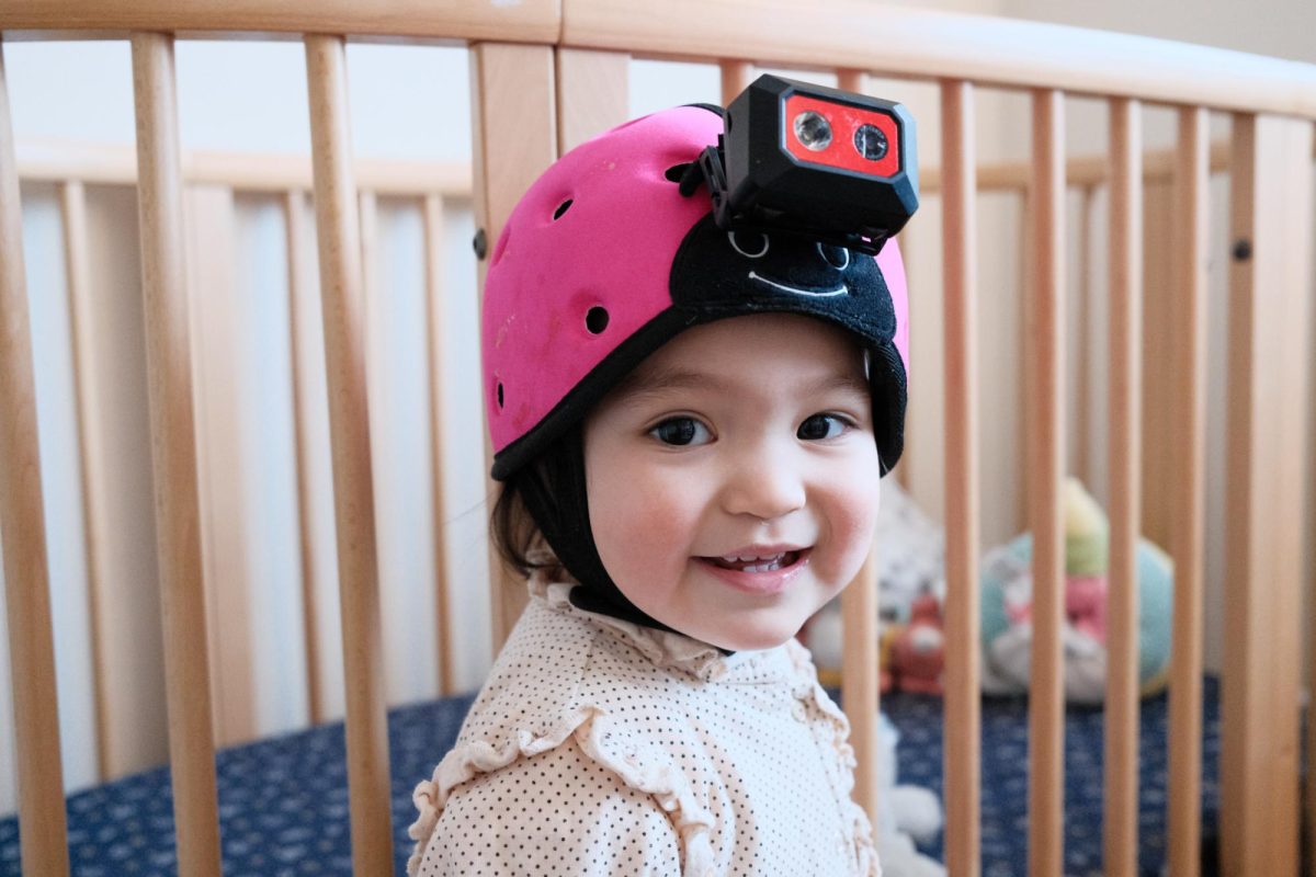 A baby wearing a head-mounted camera sits in front of a crib while smiling at the camera.