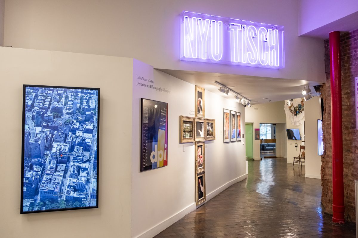 A neon purple L.E.D. sign saying “N.Y.U. TISCH” in a hallway that has white walls with several framed pieces hung on it.