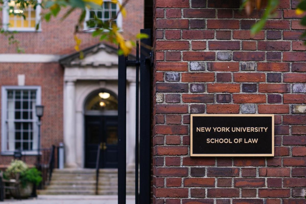 To the right there is a sign on a brick wall that reads, ‘New York University School of Law.’ Behind that wall is the entrance of a brick building.