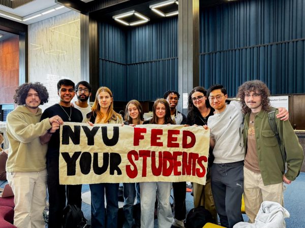 Ten students holding a banner that reads “N.Y.U. FEED YOUR STUDENTS” standing side by side in a lobby.