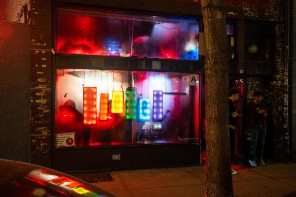 An exterior shot of a bar with “Pieces” spelled in rainbow letters on its foggy window. Colorful light is also visible through the window.