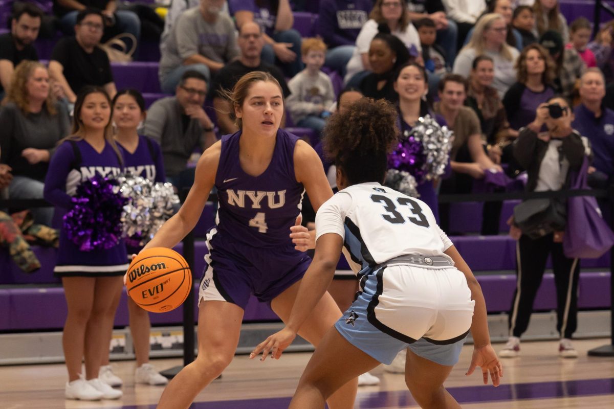 A woman wearing a purple N.Y.U. jersey dribbles a basketball in front of another woman in a white and blue jersey.