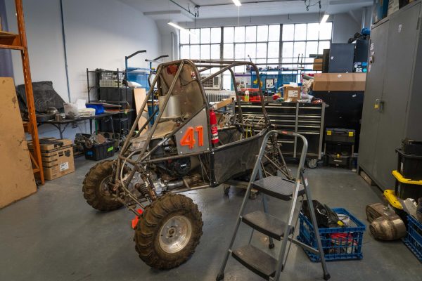 A small, metal vehicle is parked in the middle of a cluttered workshop. The vehicle has a welded cage and “41” in translucent orange letters. A fire extinguisher is visible within the car.