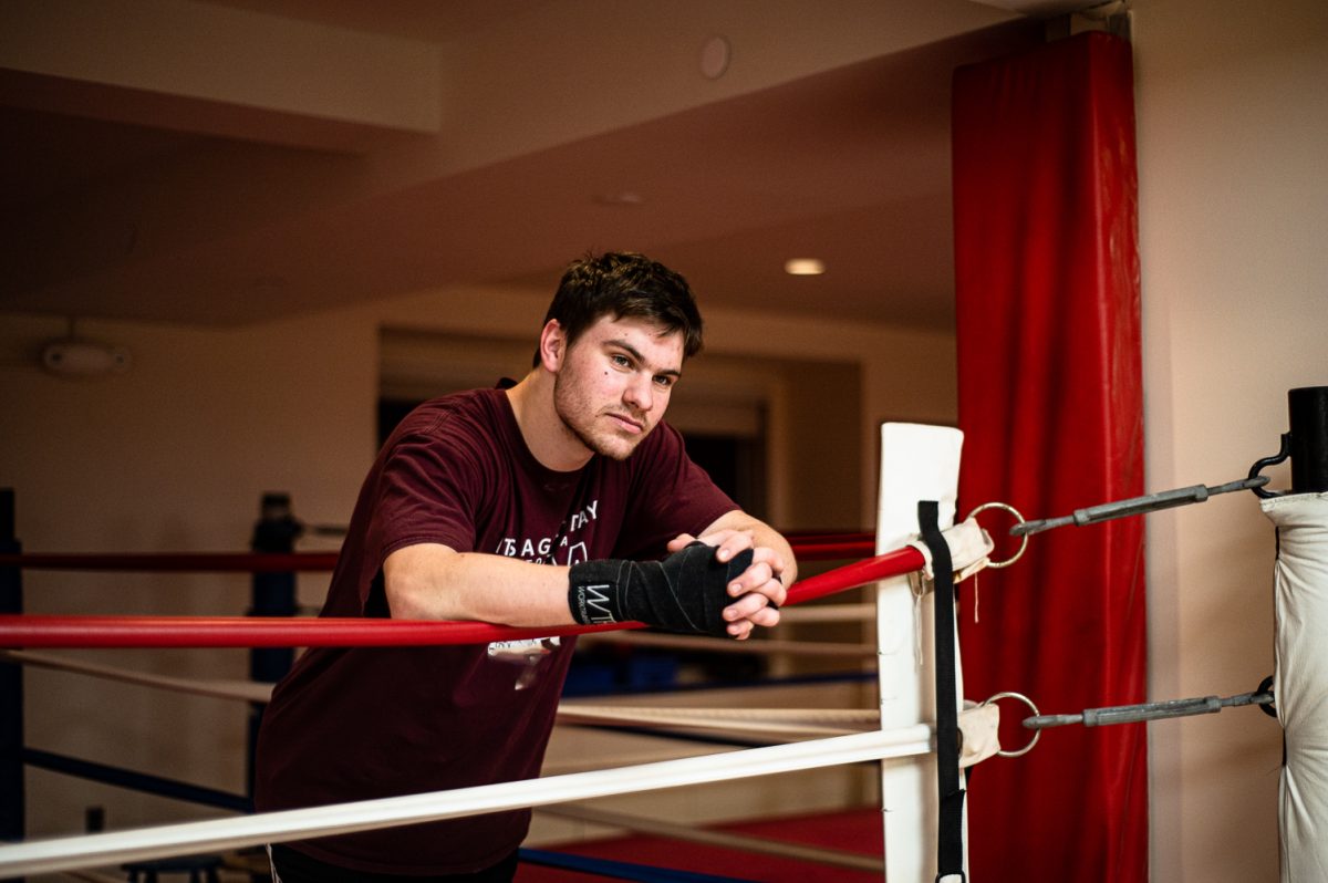 A man in a maroon shirt with white graphics leans on the plastic rope-like border of a boxing ring. He wears a wrapped, black, velcro glove on his right hand, with his hands laced together in front of him.