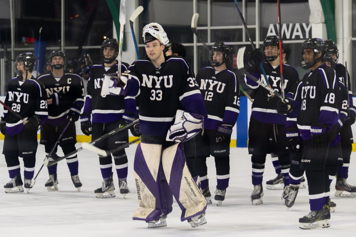 Around ten hockey players raise their sticks on the ice. Each of them are wearing their N.Y.U. black and purple uniforms. One man stands in front of them all, leading the greeting.