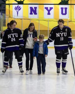 Two N.Y.U. hockey players lead an adult woman and child wearing N.Y.U. merch and shoes on the ice. The right N.Y.U. player has his hand touching the shoulder of the little boy. The woman is holding a bouquet of pink flowers.