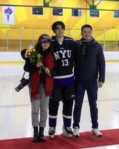 An N.Y.U. hockey player wearing the number 13 in a standard black and purple uniform poses with his parents on the ice. He is wearing his uniform while his parents wear everyday clothes and shoes. His mother is holding a bouquet of yellow flowers.