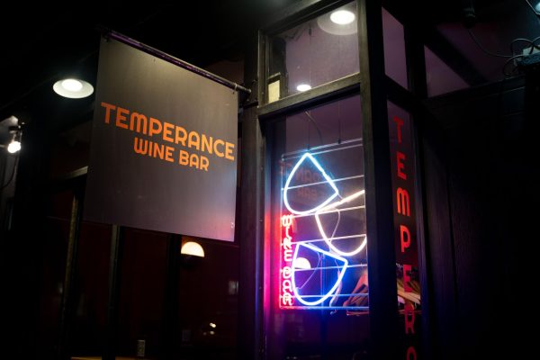 A black banner with the words “TEMPERANCE WINE BAR” on it. There is a multicolored neon sign on the side that has cups and also says “WINE BAR.”