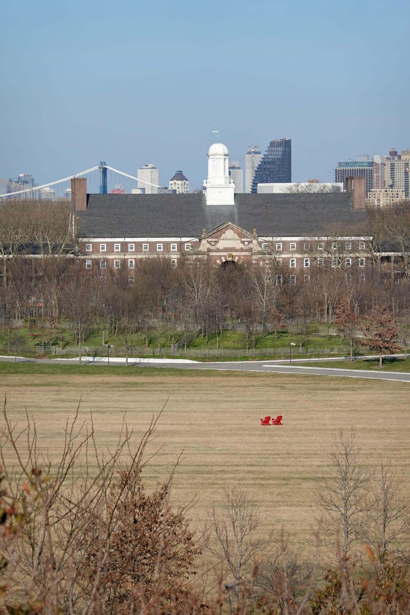 Two red chairs in an empty field with a house on Governors Island in the background.