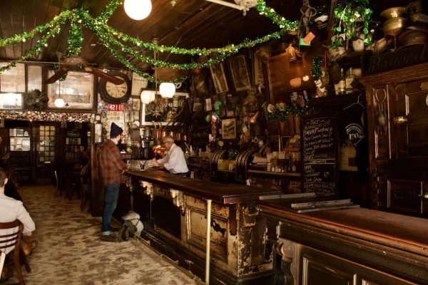 Dark wood bar interior with walls covered in various photos. Green tinsel streamers hang from the ceiling and a man in a plaid shirt orders at the bar. The older bartender wears a simple white shirt uniform. 