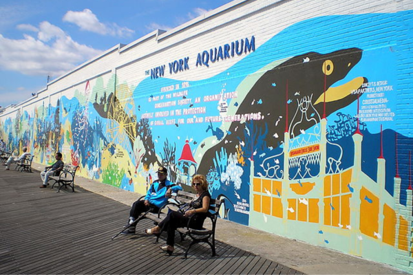A large brick wall is covered in colorful paintings of sea creatures in the ocean and raised blue letters with the words “THE NEW YORK AQUARIUM.” People sit on benches in front of the wall.