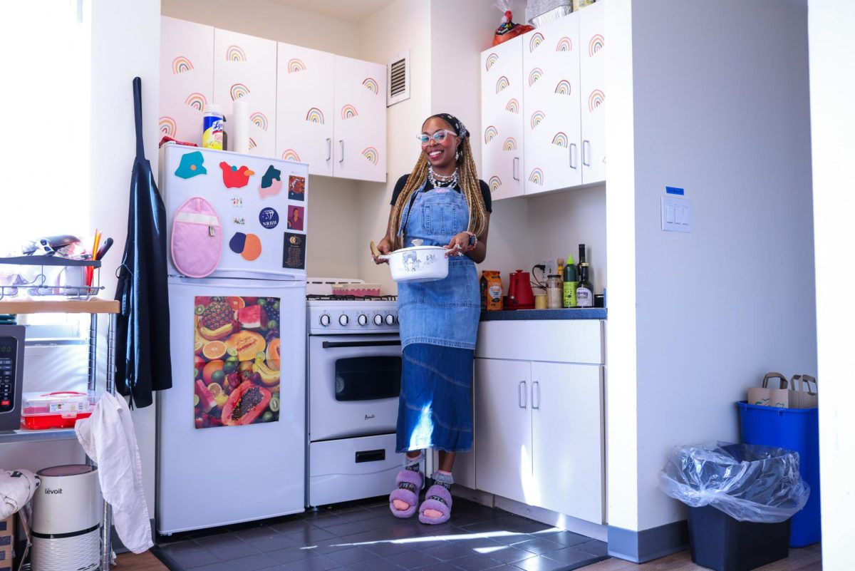 Sasha DuBose, in a blue apron and purple slippers, standing by a refrigerator and holding a white pot.