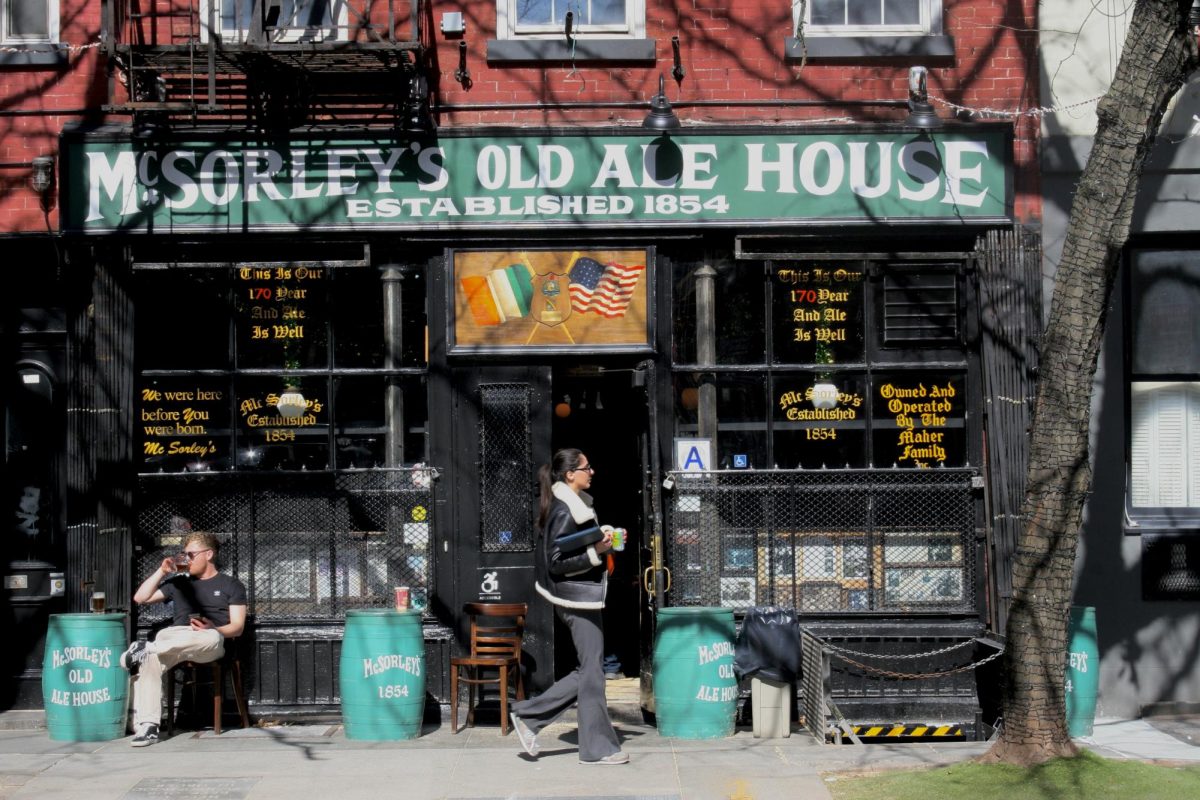 Exterior shot of a bar with a green sign that reads “MCSORLEY’S OLD ALE HOUSE ESTABLISHED 1854.” On the windows is various text in calligraphic font and four green beer barrels in front of the windows function as tables.