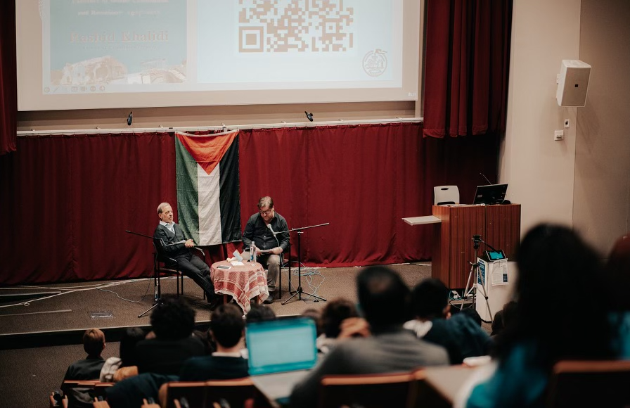 A lecture hall with students sitting in the foreground and two men sitting around a Palestinian flag.