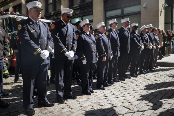 A row of officers line up shoulder-to-shoulder in white caps wear white caps and navy uniforms with a fire department patch on the sleeve.