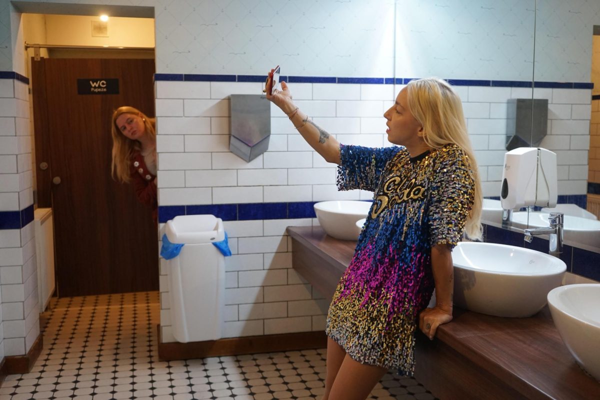 A film still of a woman leaning on a bathroom sink and taking a picture of herself. To the left, a woman is peaking her head into the bathroom.