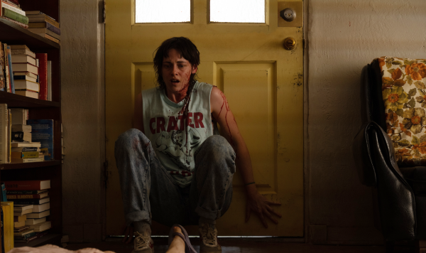 A woman with a shocked expression and covered in blood crouches in front of a door. She’s staring at someone lying on the floor out of frame with their feet exposed.
