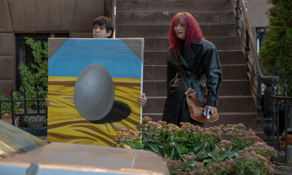 Two people standing outside. The person on the left is holding a large canvas with an egg painted on it.