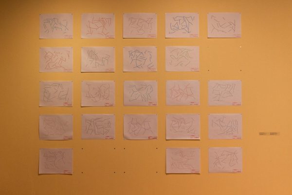 A yellow wall with 22 sheets of white paper, each with drawings of short, scattered lines.