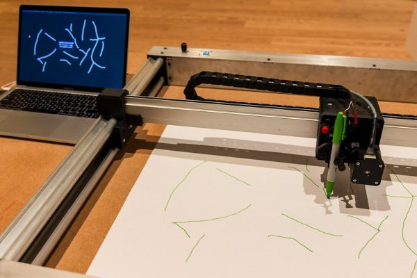 A machine uses a green sharpie to draw on a sheet of white paper. Next to it, a laptop displays the pattern being copied.