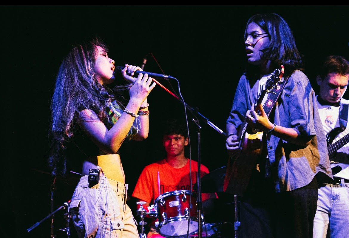 A band performs on stage, a woman sings into the mic while two men play guitar and drums beside her.