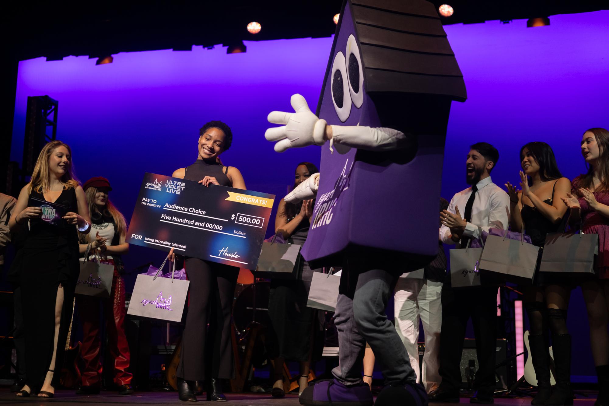 A purple house mascot gestures toward a woman holding a giant cardboard check for $500.