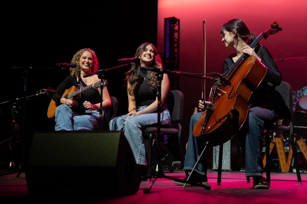 Three women perform on stage. The far right woman holds a cello while the woman on the far left has an acoustic guitar.