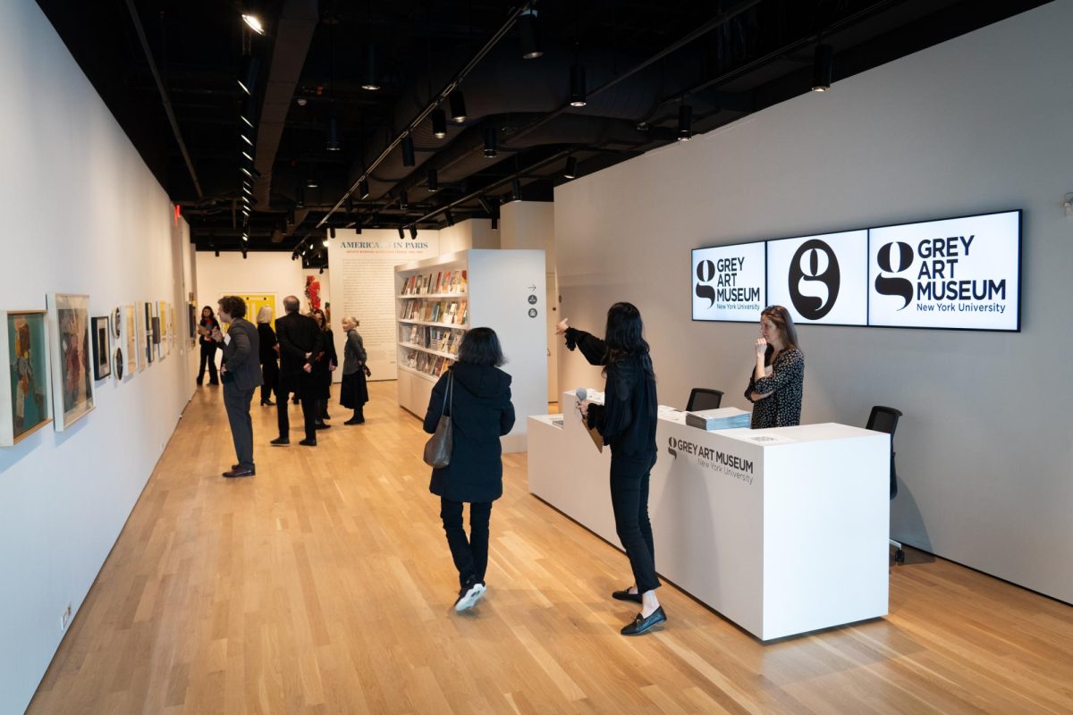 The interior of a gallery where a white front desk with a logo saying “GREY ART MUSEUM” printed on it is placed near the entrance. A group of people is looking at photographs and paintings displayed on white walls.