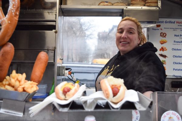 A woman in a black hoodie stands inside of a food cart behind hotdogs and a salted pretzel.